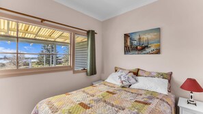 Een slaapkamer met een groot raam, een bed met quilt, een nachtkastje, een rode lamp en een schilderij aan de muur.