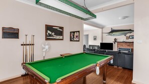 Une table de billard recouverte d’un drap vert, un porte-queues et un ventilateur.