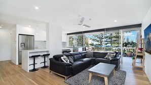 Un espacio habitable moderno con un sofá de cuero negro, una mesita, una zona de cocina y un balcón con plantas en macetas.