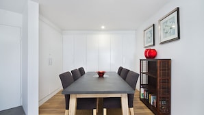Un salón de conferencias con una mesa larga, seis sillas, un jarrón rojo y una estantería llena de libros.