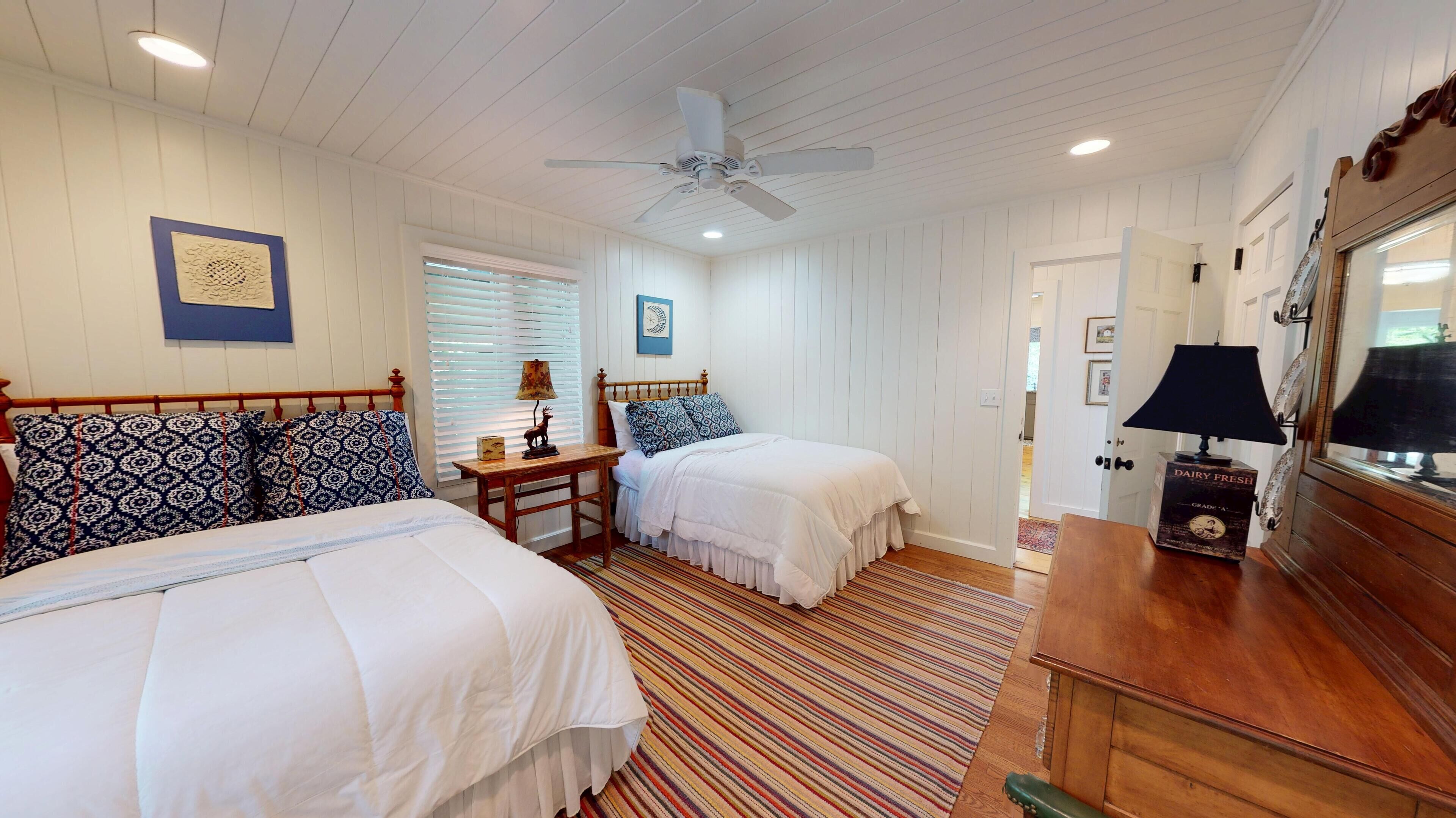 Une chambre avec deux lits, un ventilateur de plafond, une commode en bois et un tapis à rayures.