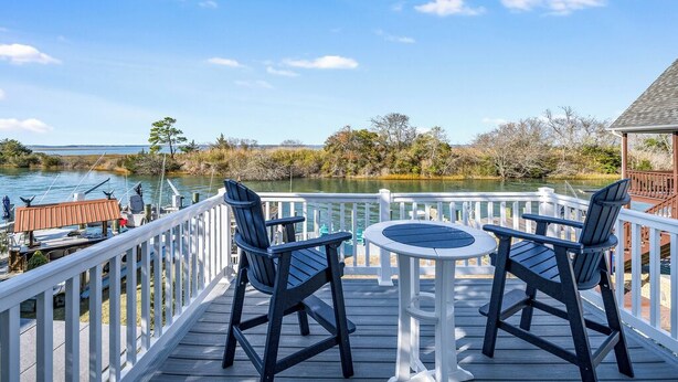 A deck with two Adirondack chairs and a small round table, overlooking a calm lake with boats and a dock in the background. - Family Tides by Seaside Vacations & Sales (Chincoteague Island)