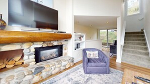 A modern living room with a stone fireplace, a flat-screen TV, a comfortable armchair, and a staircase leading to an upper level. - Knob Hill Ridge Townhome 1 - Modern Decor Right in Downtown and Pet Friendly (Ketchum)