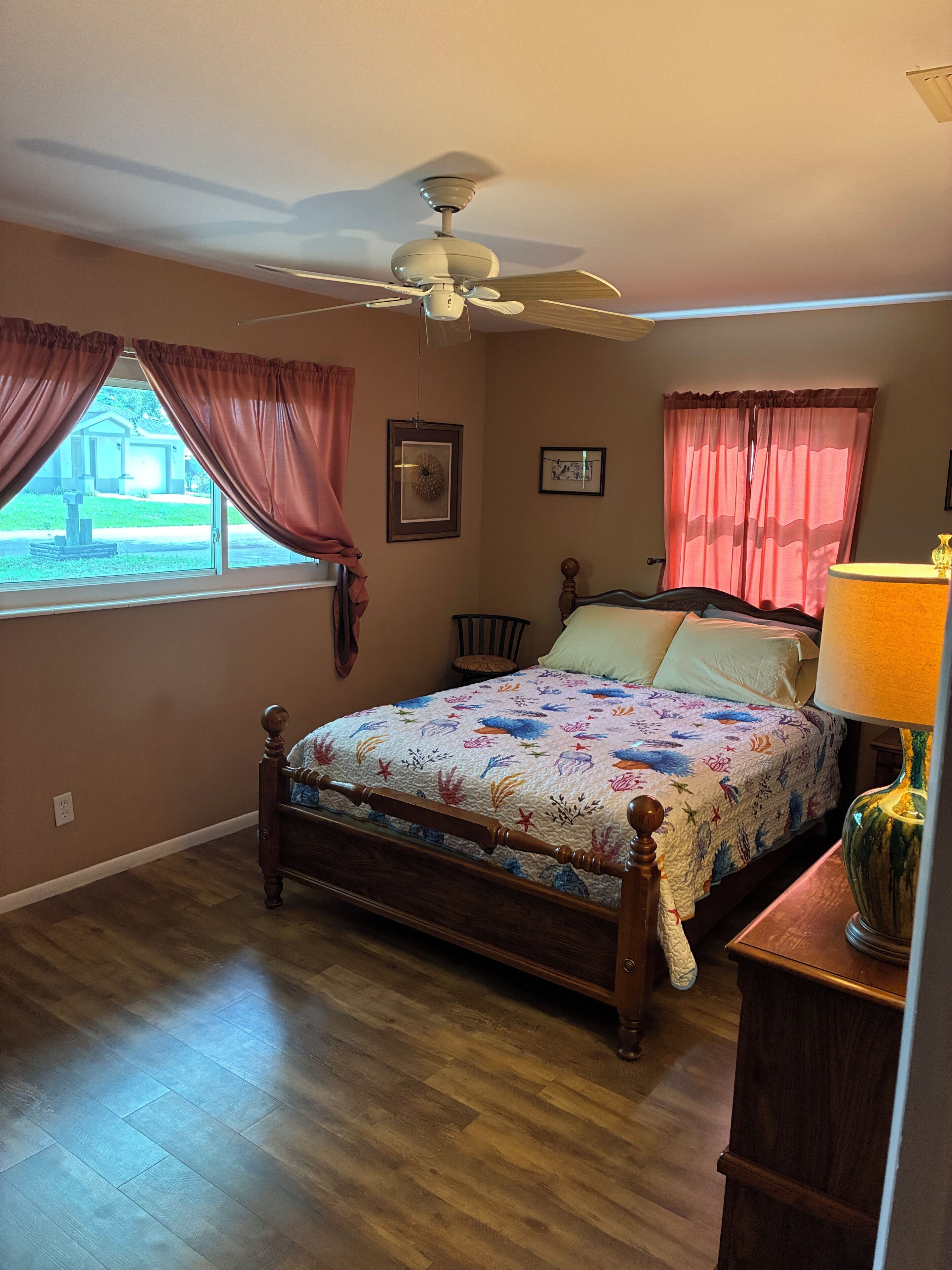 Un dormitorio con cama, ventilador de techo, ventana con cortinas, una silla, mesita de noche con lámpara y un cuadro en la pared.