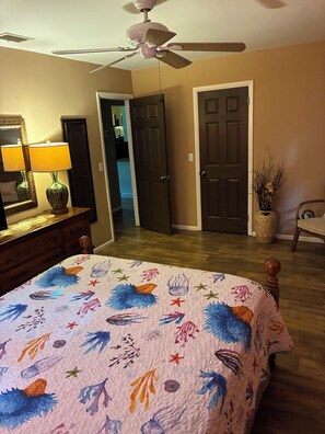 A bedroom with a bed, a ceiling fan, a dresser, a lamp, and a door leading to another room.