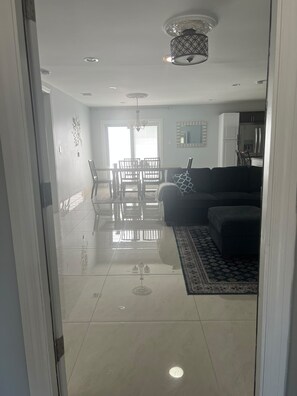 A modern living room with a glass door entrance, a dining area with a chandelier, and a kitchen with white cabinets.