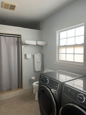 A laundry room with a front-loading washer and dryer, a toilet, and a shower area.