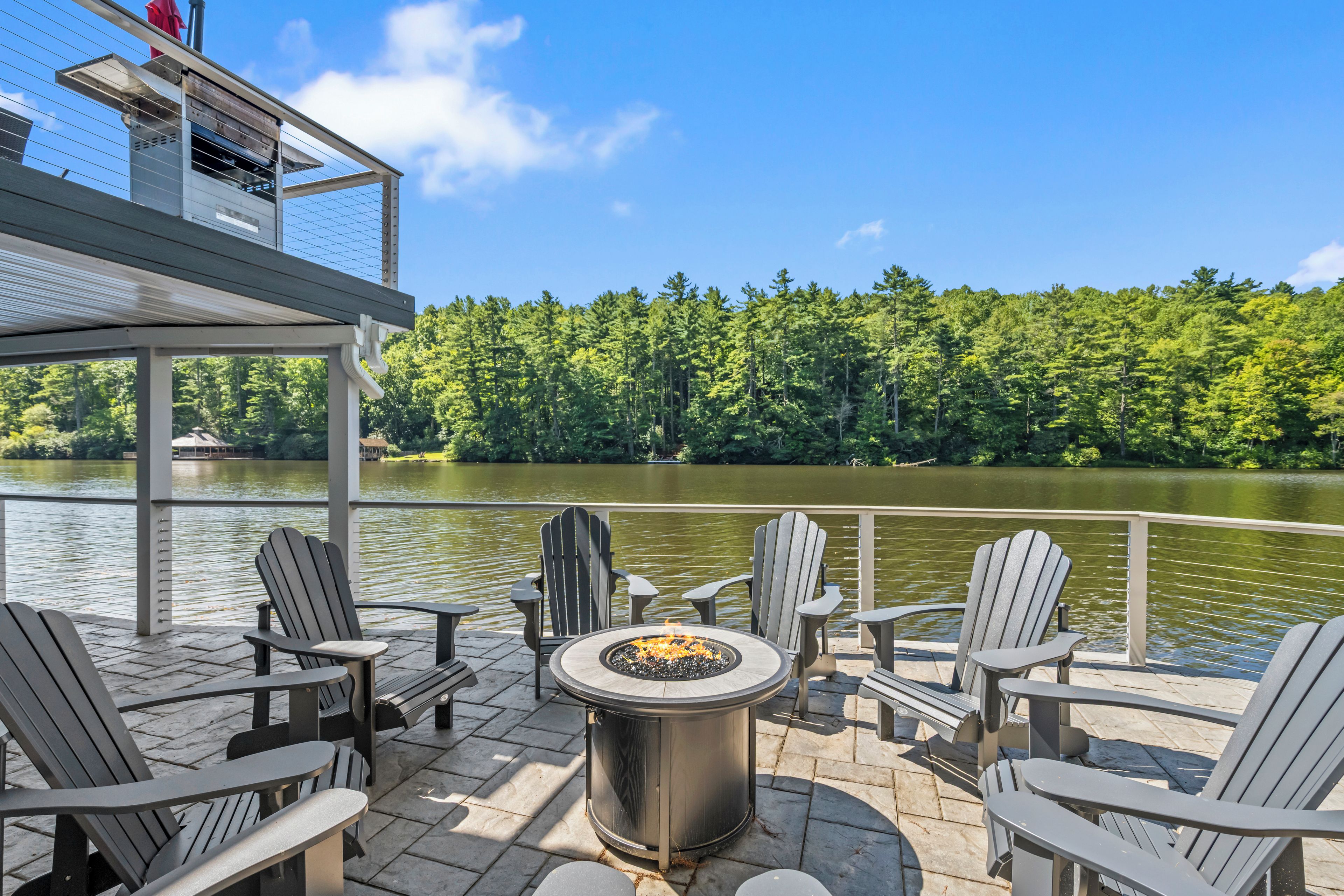 A deck with Adirondack chairs and a fire pit overlooking a lake, with a forested shoreline in the background.