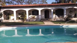 Un área de piscina con una pileta de baldosas, un edificio blanco con arcos y un techo de tejas rojas.