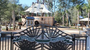 Una puerta con letrero que dice 'Adventure Playground', una estructura de juego de madera y un quiosco de información del parque.