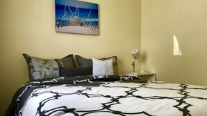 A neatly made bed with a patterned bedspread, a nightstand with a lamp, and a painting of a beach scene on the wall. - Relaxing beach vacation - beautiful, quiet and clean (Cooks Beach)