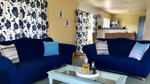 A living room with two dark blue sofas, a glass coffee table with a tray of drinks, and a kitchen in the background. - Relaxing beach vacation - beautiful, quiet and clean (Cooks Beach)
