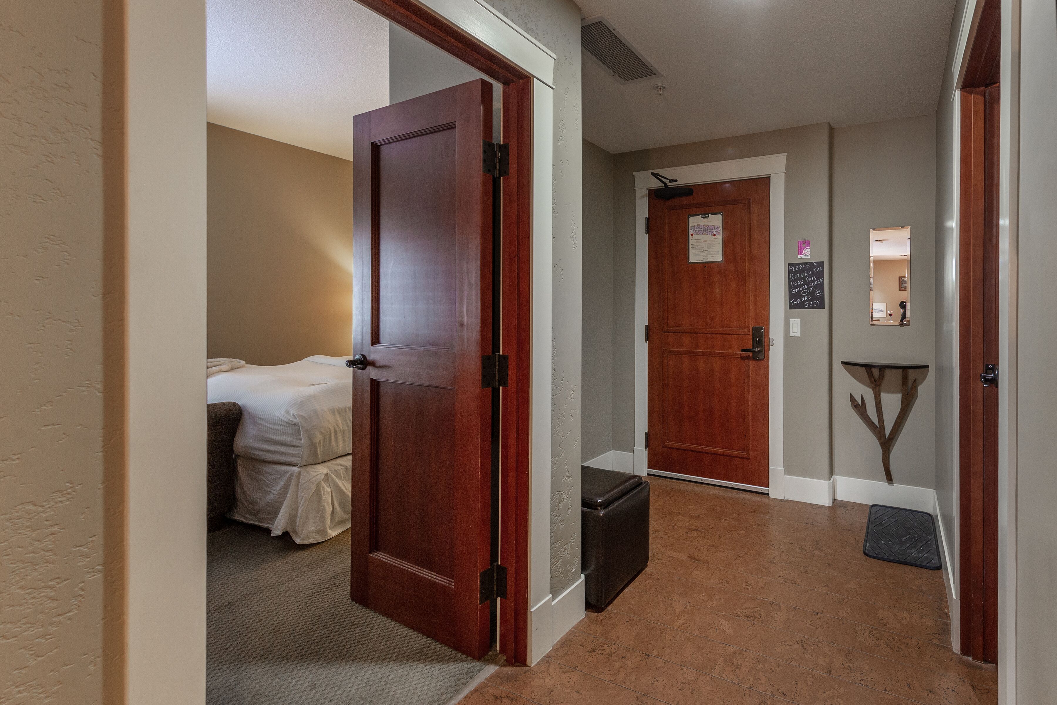 Un couloir d’hôtel avec une porte en bois, un lit, une valise et une petite table avec une chaise.