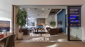 A hotel lobby with a 24-hour front desk, check-in area, and a large potted plant.