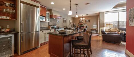 A modern kitchen with a large island, stainless steel appliances, wooden cabinets, and a dining area with a sofa and table.