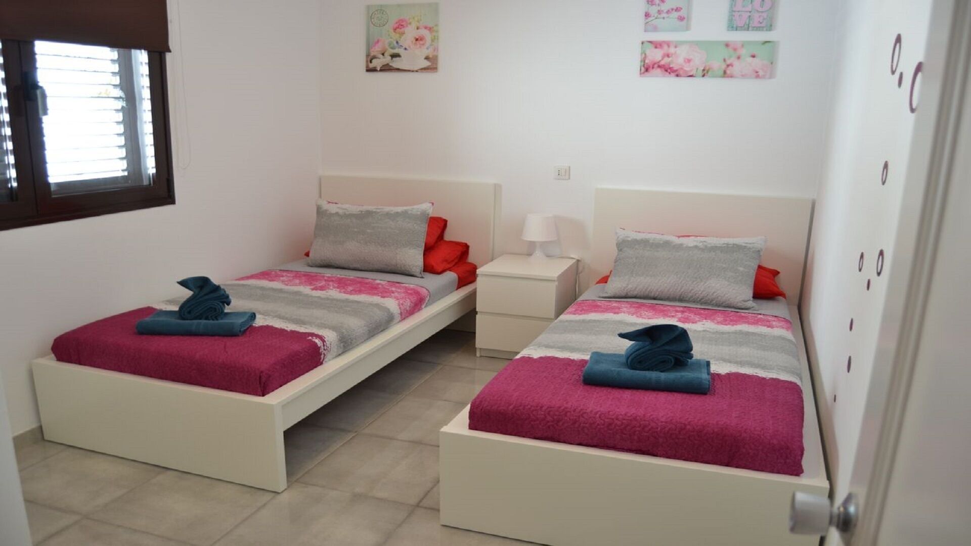 Habitación doble con dos camas, cada una con una manta rosa y gris, una mesita de noche blanca y una ventana con persianas.