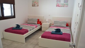 Quarto duplo com duas camas, cada uma com cobertor rosa e cinza, uma mesinha de cabeceira branca e uma janela com persianas.