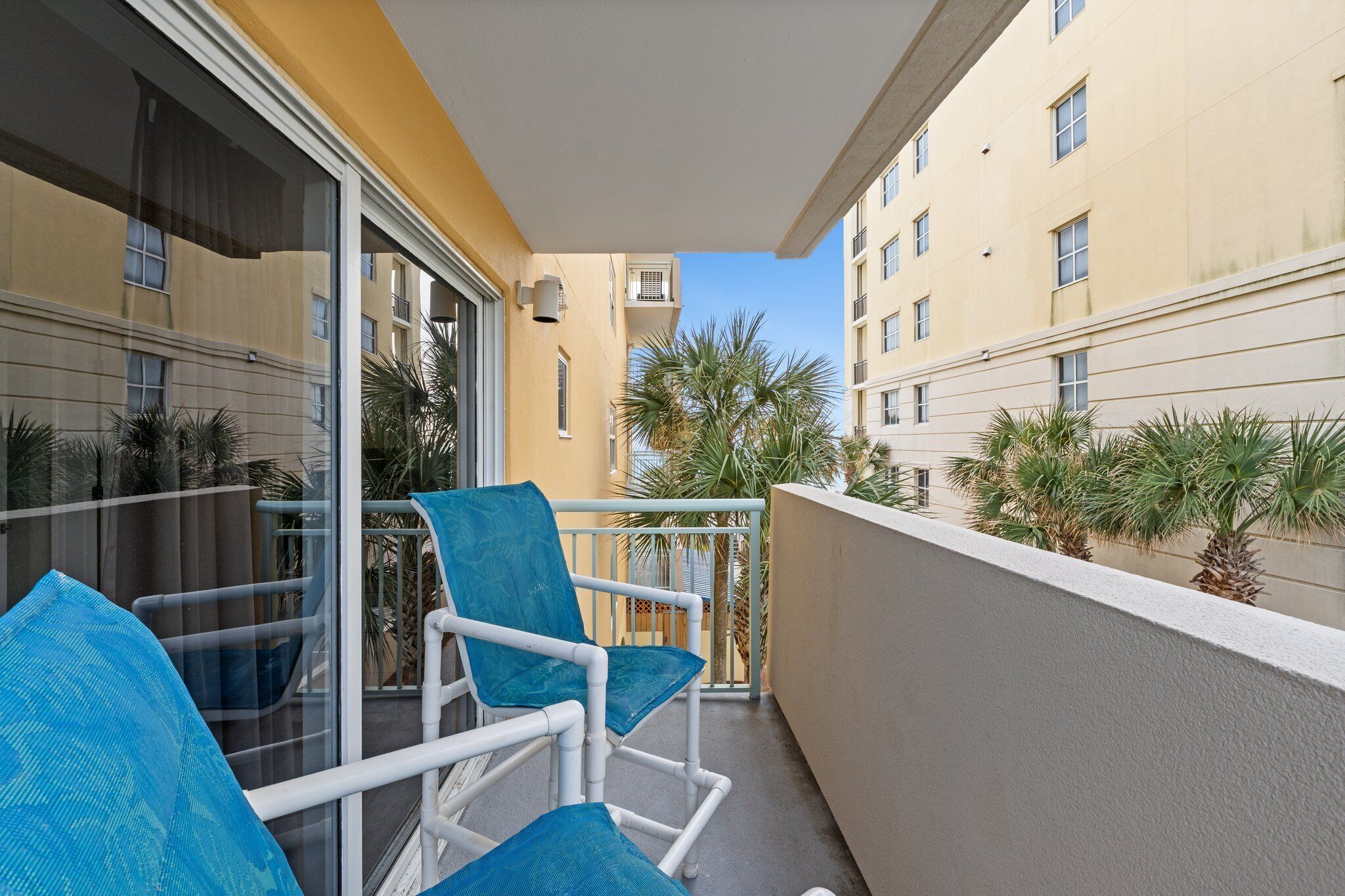 A balcony with a white chair and a blue cushion, a glass door, and a view of palm trees and a building.