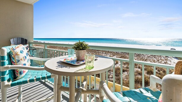 En balkon med udsigt til stranden, et bord med en plante og et glas, samt en stol med pude.