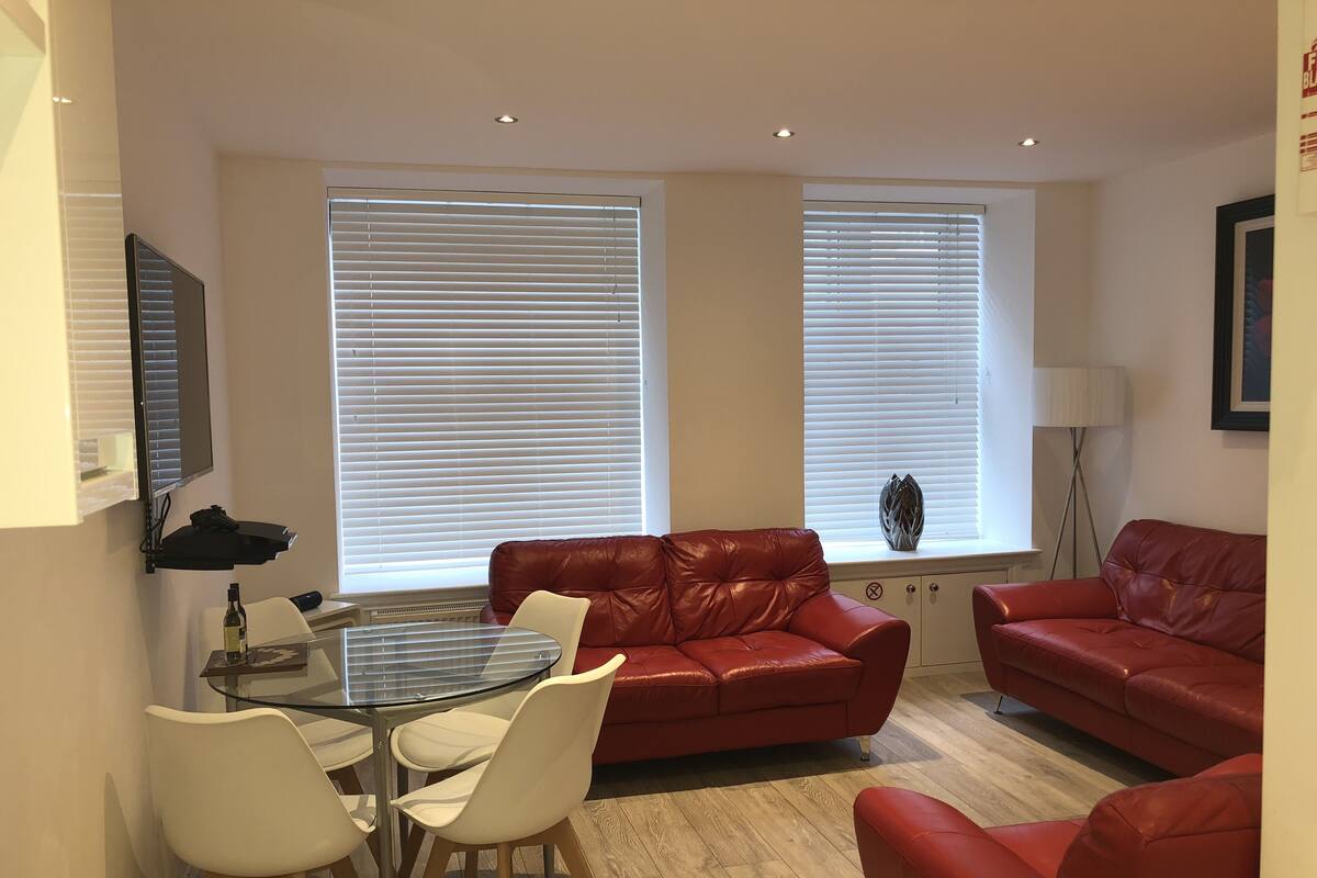 A living room with a red leather sofa, a glass coffee table, and white chairs. There are two windows with blinds, a floor lamp, and a framed picture on the wall.