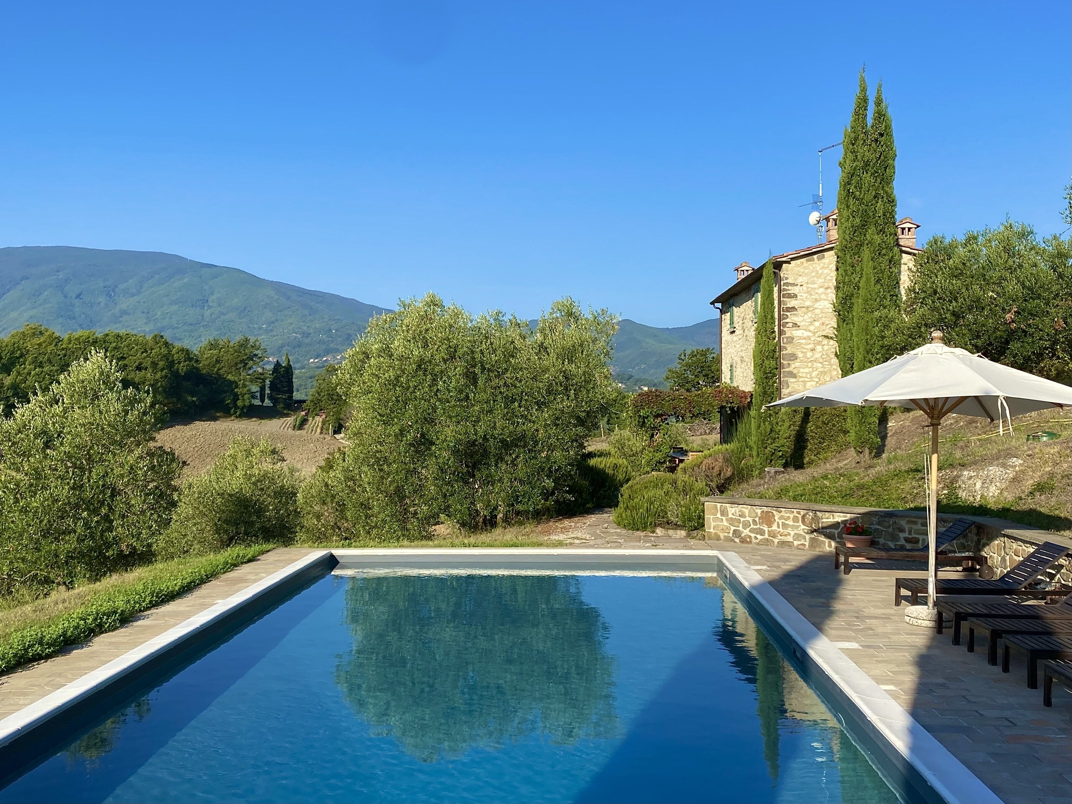 A swimming pool with a stone border, surrounded by greenery and a stone building with a white umbrella.