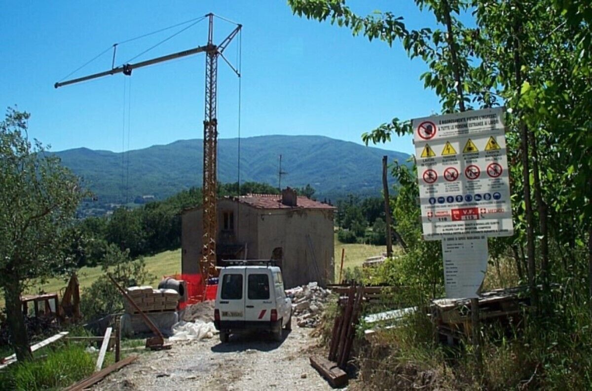 Un chantier de construction avec un fourgon blanc, une grue et un petit bâtiment. On y voit un panneau avec des symboles d’avertissement et du texte.