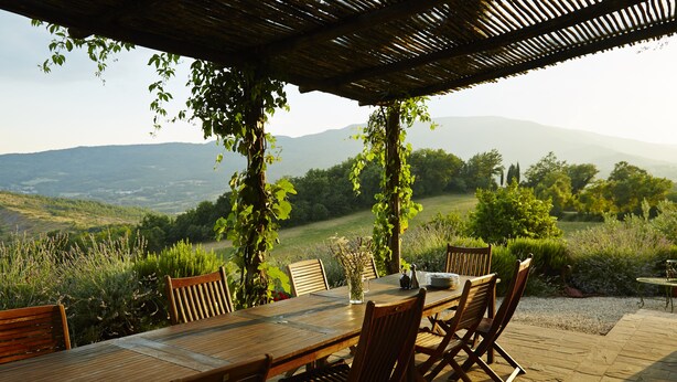 Un área de comedor al aire libre con una mesa y sillas de madera, rodeada de follaje y vistas a colinas distantes.