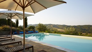 Zona junto a la piscina con sillones de descanso y sombrillas, rodeada de un muro de piedra y vegetación, con vistas a un paisaje montañoso.