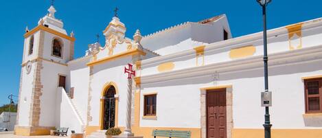 A white building with a bell tower and a cross, a bench, and a street lamp.