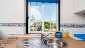 Cozinha com pia de aço inoxidável, janela com cortina azul e vista para o verde lá fora.