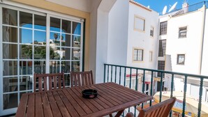 Mesa de madeira com cadeiras numa varanda, com porta de vidro e vista para edifícios vizinhos.