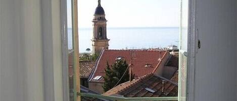 A room with a window overlooking a coastal town, featuring a bell tower and terracotta rooftops.
