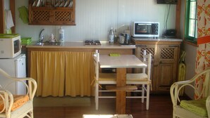 A compact kitchen with a dining area, featuring a wooden table, chairs, a microwave, a sink, and a window with floral curtains.