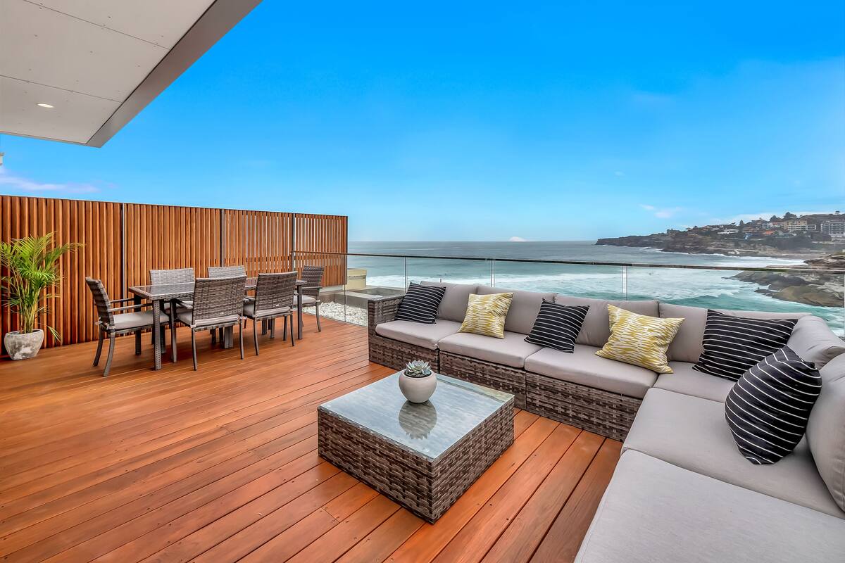 A modern outdoor patio with a wooden deck, a dining area with wicker furniture, a cushioned seating area with patterned cushions, and a view of the ocean and coastal town.