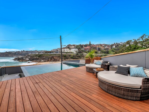 Una terraza de madera con piscina, mobiliario de mimbre y vistas al océano y casas en la colina.