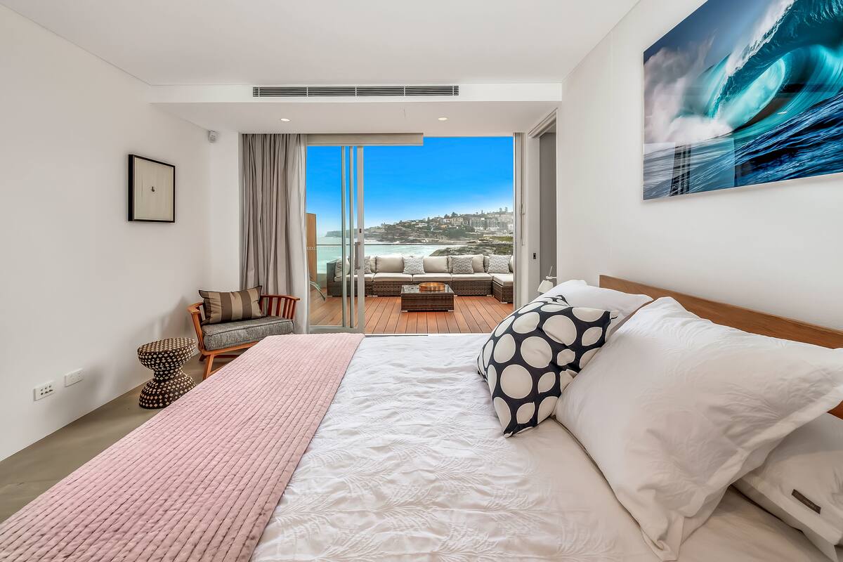 A modern bedroom with a large bed, a view of the ocean, and a balcony with outdoor seating.