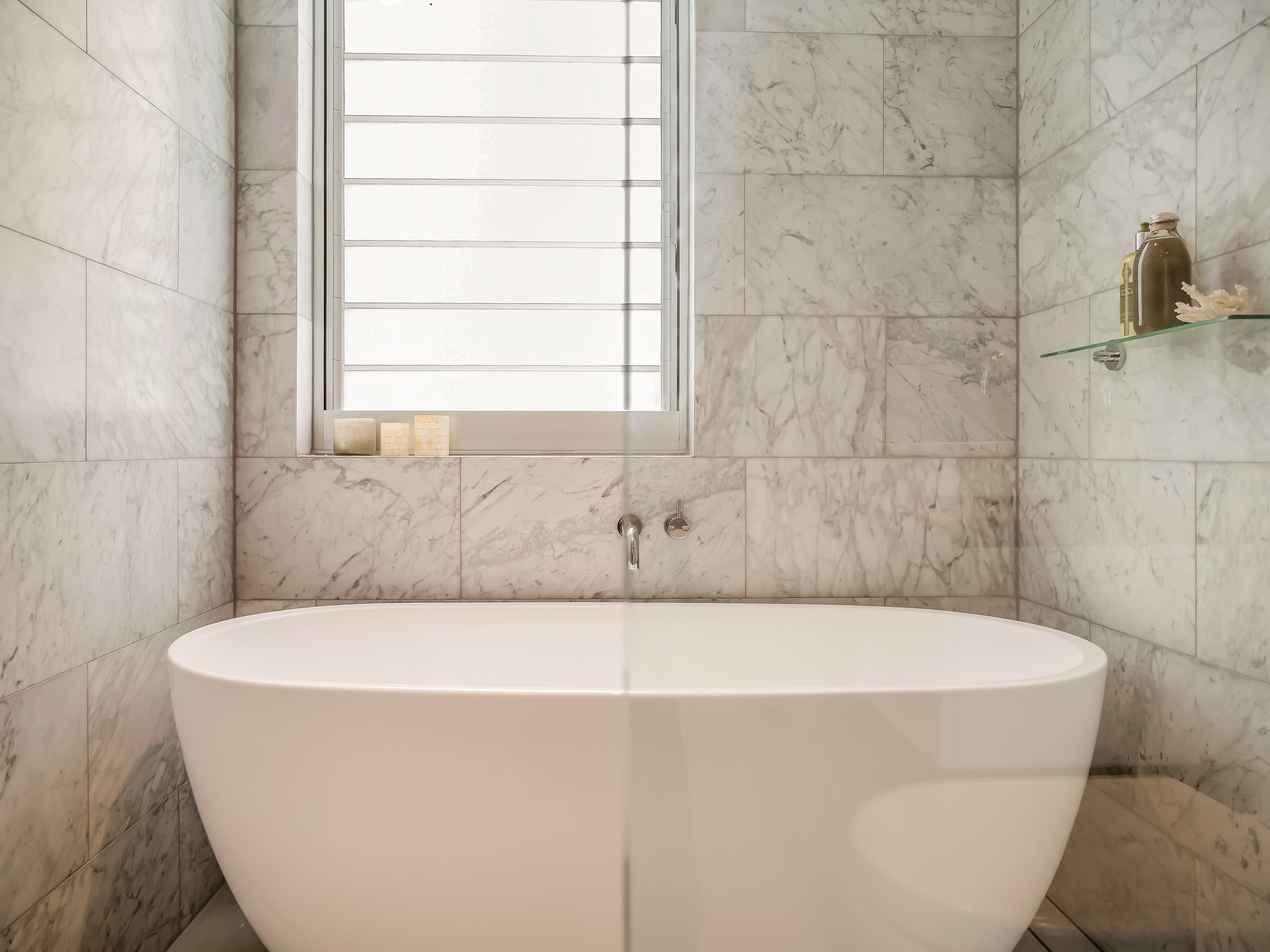 Un baño moderno con una bañera independiente, paredes de mármol, una ventana y un estante de vidrio con artículos de aseo.