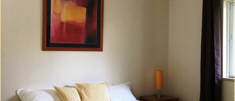 A double bed with white linens, a bedside table with a lamp, a framed abstract painting on the wall, and a window with curtains.