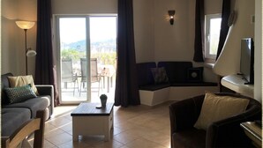 Un salon comprenant un canapé, un fauteuil et une petite table. Une porte-fenêtre coulissante donne sur un espace extérieur équipé d’une table et de chaises. On peut voir une lampe et un luminaire fixé au mur.