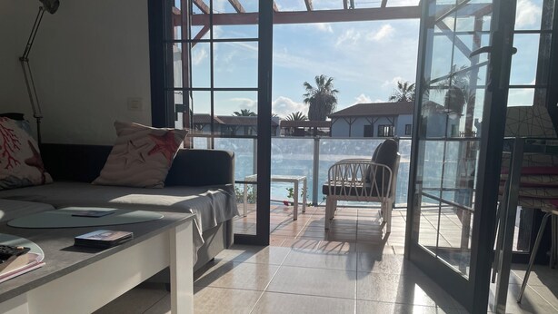 A modern living room with a sofa, coffee table, and a view of a pool and palm trees through large windows. - Relaxing, Poolside bungalow, 1 Bedroom luxury modern,  Sleeps 4 (Caleta de Fuste)