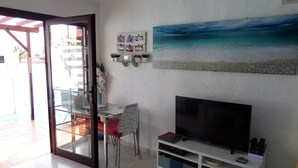A living room with a TV, a glass table, chairs, a bookshelf, and a painting of a beach. - Relaxing, Poolside bungalow, 1 Bedroom luxury modern,  Sleeps 4 (Caleta de Fuste)