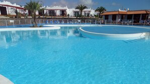 A clear blue swimming pool with a curved design, surrounded by a low stone wall and palm trees. - Relaxing, Poolside bungalow, 1 Bedroom luxury modern,  Sleeps 4 (Caleta de Fuste)