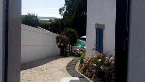 Un chemin mène à un espace piscine agrémenté d’un mur blanc, d’une porte bleue et de plantes en pot.