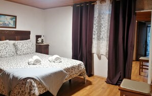 A neatly made bed with patterned bedding and rolled towels, a small bedside table with a lamp, and a view of a room with wooden flooring and a door.
