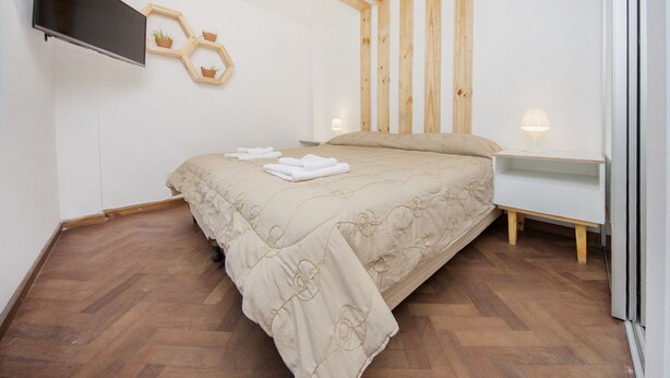 Een slaapkamer met een bed, een aan de muur bevestigde televisie, een nachtkastje met een lamp, en houten balken aan het plafond.