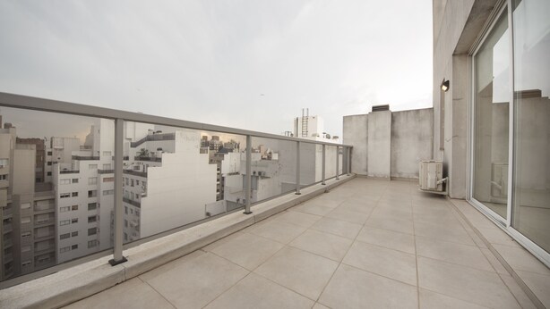 Uma varanda com guarda-corpo de vidro, piso de azulejos e vista para prédios da cidade.