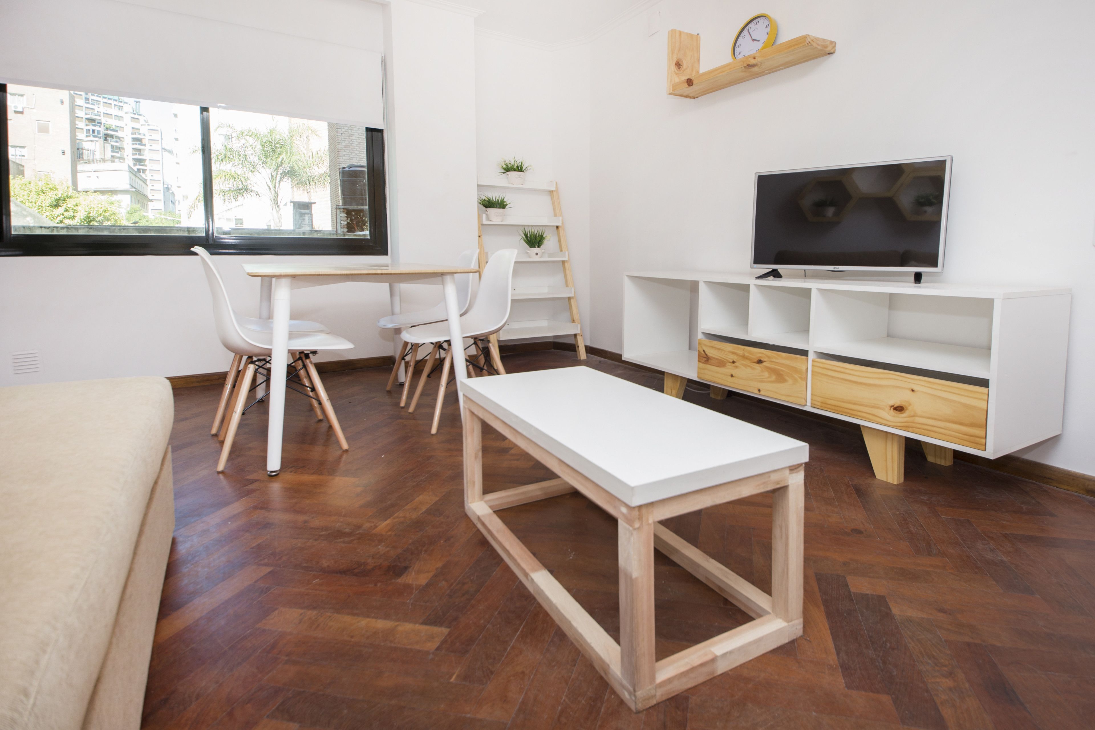 A modern living room with a wooden floor, white furniture, a TV on a wooden stand, and a window with a view of buildings.