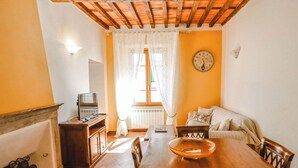 Un coin repas avec une table en bois, des chaises, un téléviseur posé sur un meuble, une horloge au mur et une fenêtre avec des rideaux.