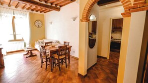 Un coin repas avec une table et des chaises en bois, une horloge au mur et une cuisine visible à travers une arche.