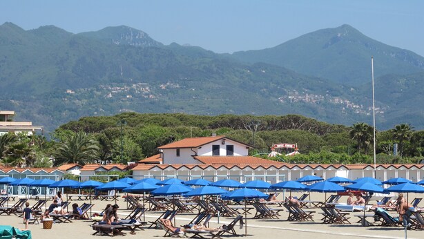 Une plage bordée de rangées de chaises longues et d’ombrelles bleues, un bâtiment avec un toit rouge, et en arrière-plan, des collines verdoyantes.
